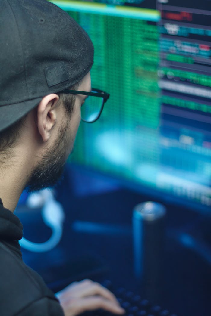 A man with glasses and a cap focused on computer programming in a tech environment.